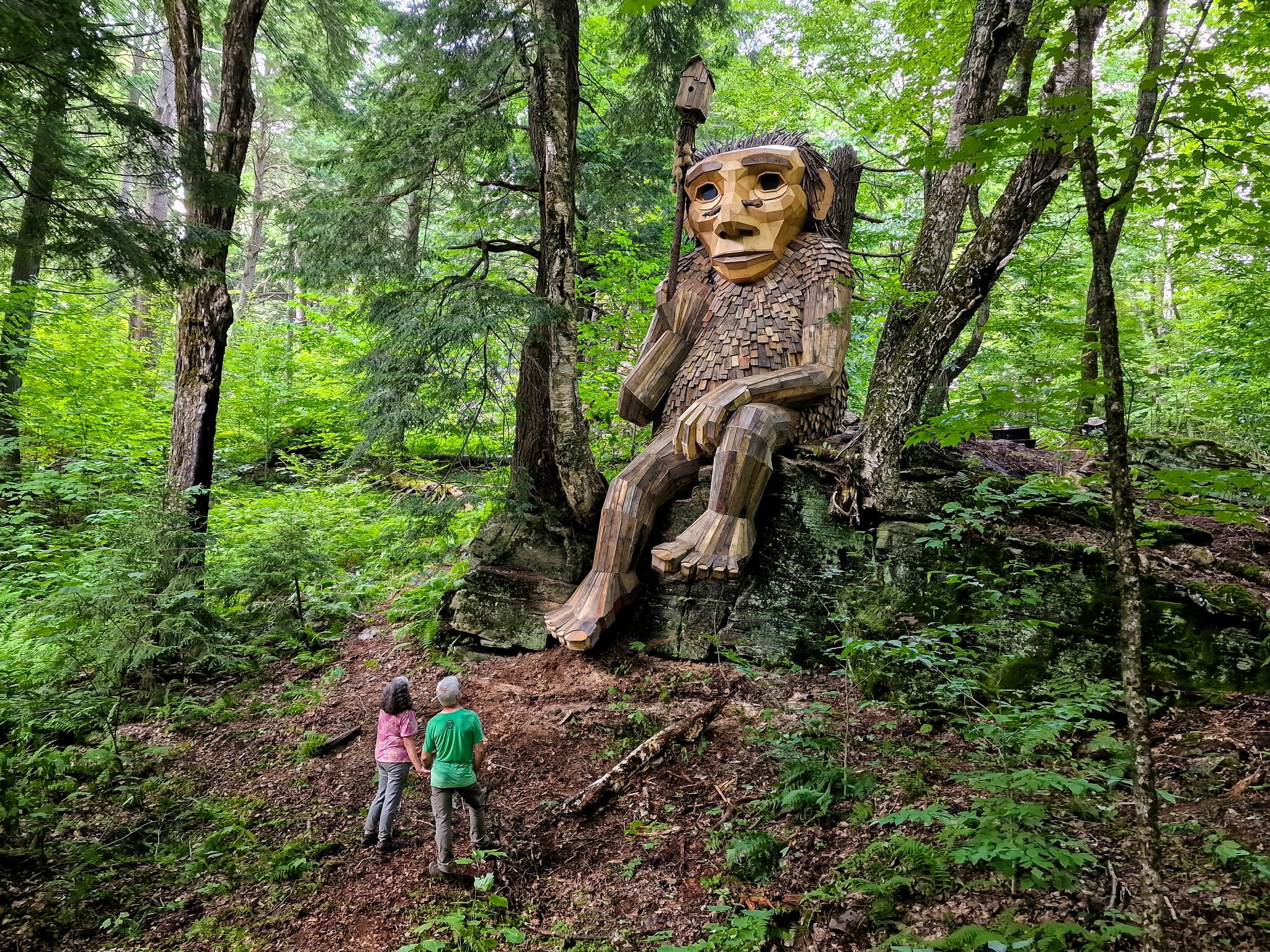 A giant wooden troll sculpture by Danish artist Thomas Dambo sits on a rock in a Vermont wooded area, while two people look at it.