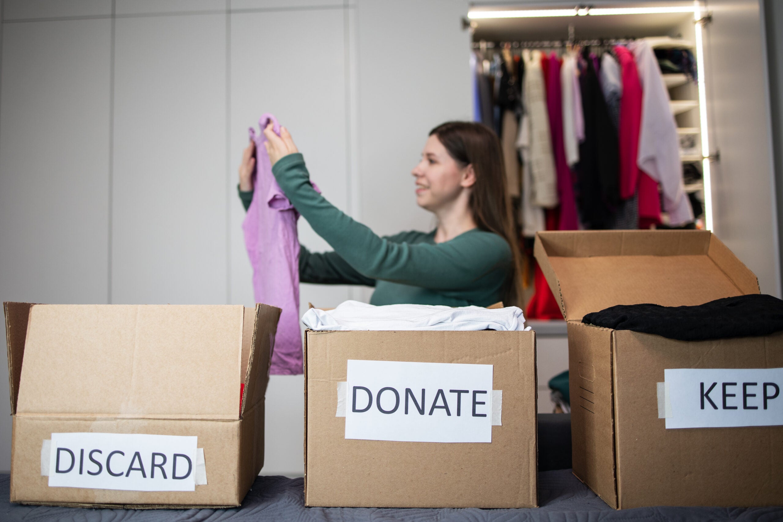 Unrecognizable woman sorts through her wardrobe. Three craft cardboard boxes of keep, discard, donation, donate. Charity clothing donations, help low income families, recycling, declutter, sustainable